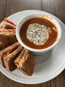 Picture of tomato soup in a white ramiken dish. It is topped with cottage cheese and dried basil. On the side is a grilled cheese which includes melted cheese and pepperoni slices. The grilled cheese and soup are on a white plate.
